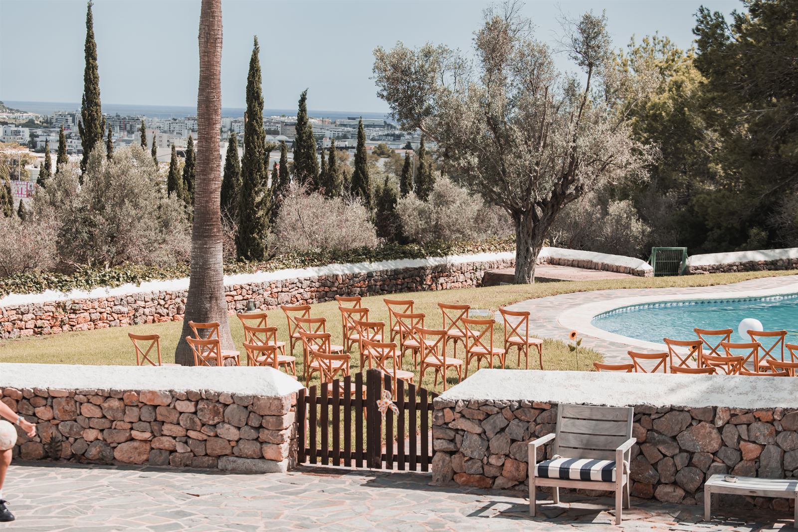 Wooden chairs set out on the lawn for a ceremony, with the pool and olive trees behind